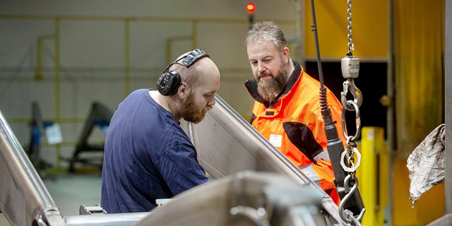 Robert Wallgren, RSO i orange arbetskläder tittar på när en arbetskamrat i blå t-shirt genomför ett arbetsmoment.