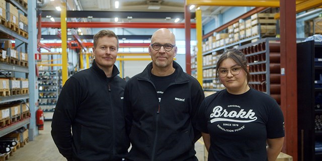 Tre personer i svarta tröjor står på ett lager, tittar mot kameran.