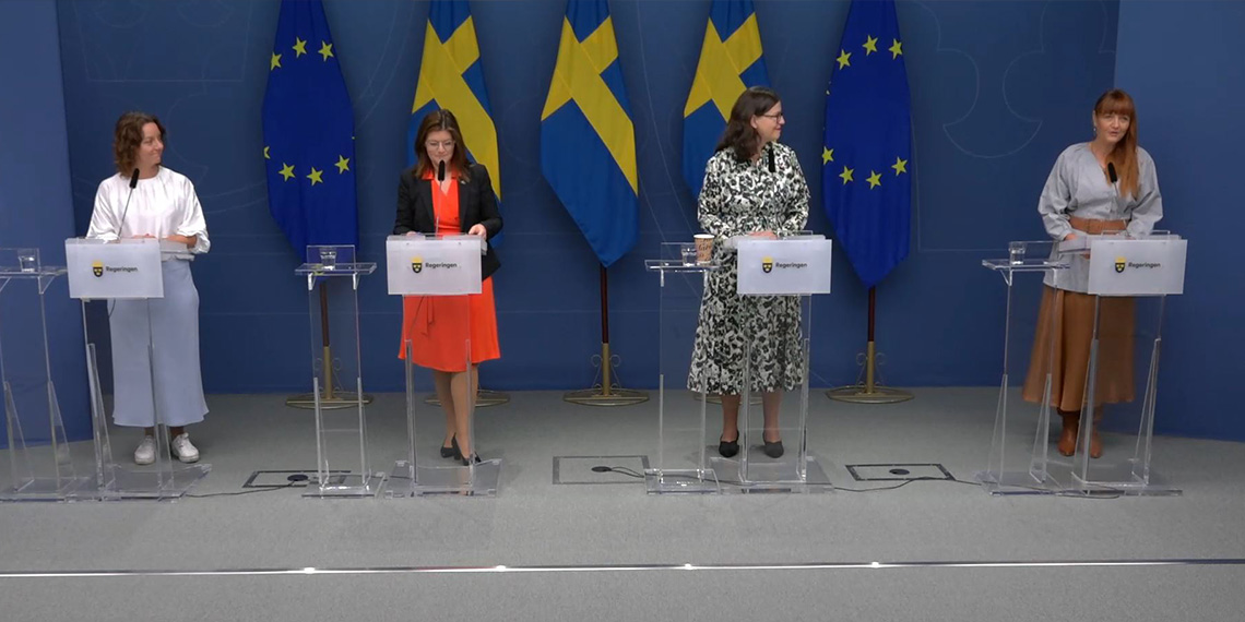IF Metalls förbundsordförande deltar vid regeringens pressträff. Fyra personer i talarstolar med svenska flaggan i fonden.