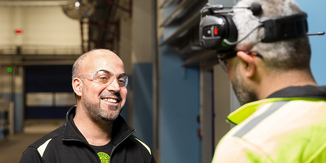 Två personer står och pratar med varandra på en arbetsplats. En har skyddsombudsmärket på tröjan och ler.