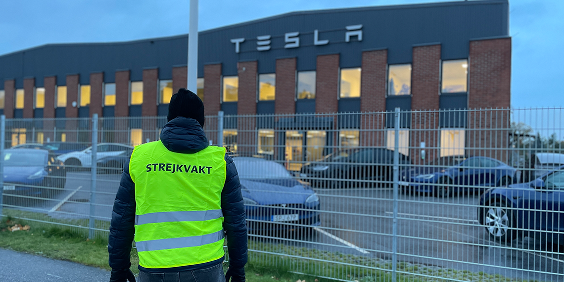Ryggen på en strejkvakt syns framför ett hus som det står Tesla på