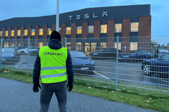 En person med varselväst står med ryggen mot kameran och tittar mot Teslas verkstad i Segeltorp. På ryggen står texten "Strejkvakt"