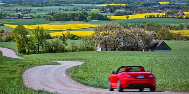 Röd bil kör på slingrande väg genom sommarlandskap.