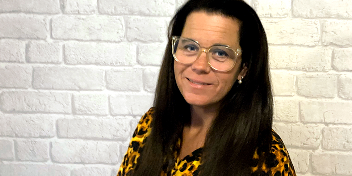 Bild på Anna Björklund som står framför en vit tegelvägg. Anna ler.