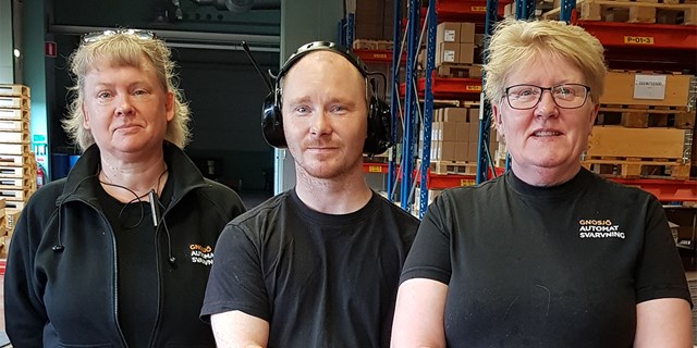 Tre personer i svarta arbetskläder märkta med företagsnamnet Gnosjö automatsvarvning i lagermiljö.