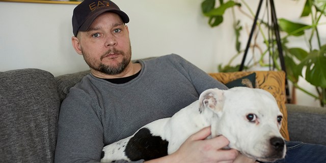 Kent Dahlin i grå soffa med en stor, svart-vit hund i famnen.