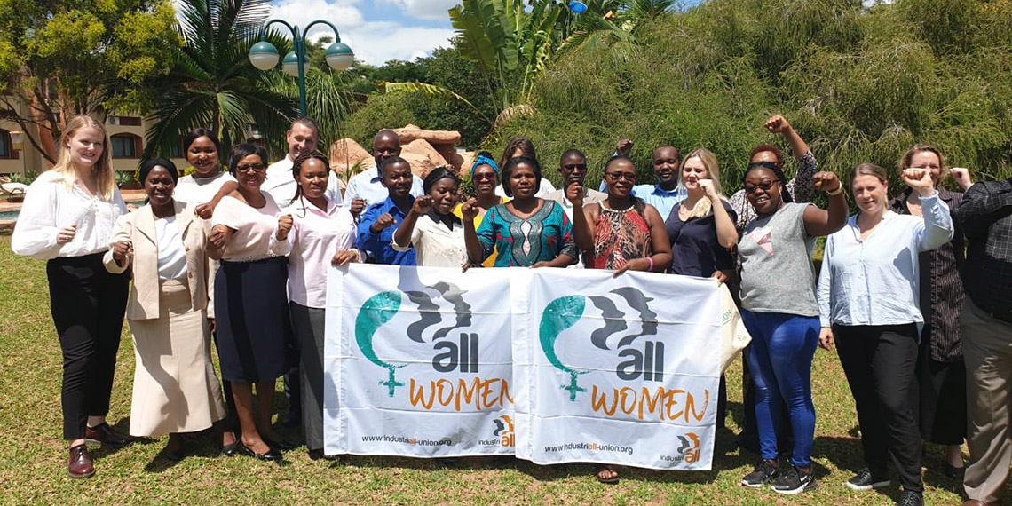 En grupp kvinnor i grön omgivning med träd och palmer håller upp banderoller där det står "All women".