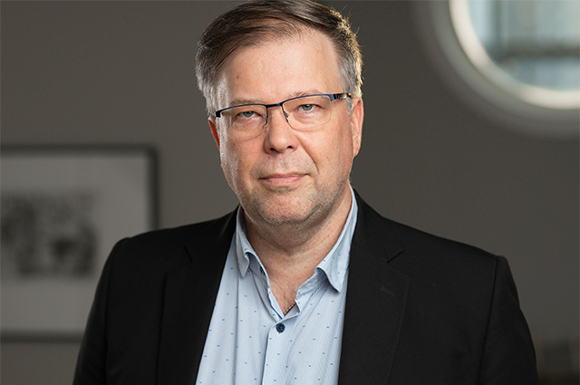 Porträtt av person i kavaj och blå skjorta mot bakgrund i skugga.