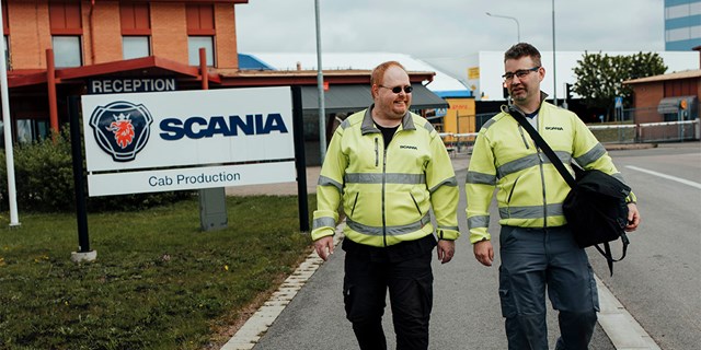 Två personer i arbetskläder till fots utomhus, intill en stor skylt med texten "Scania".