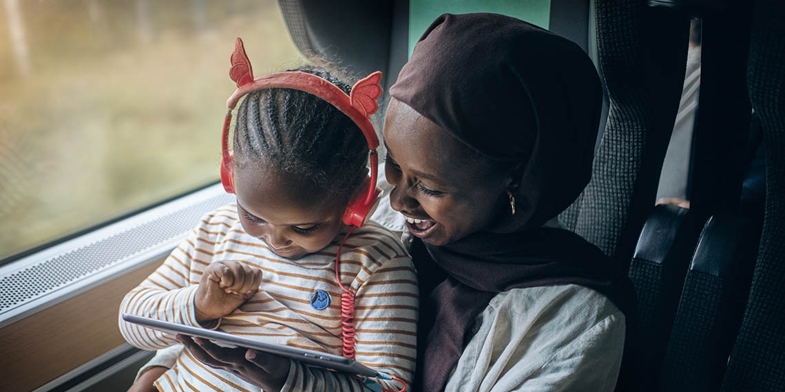 En kvinna och ett barn sitter på ett tåg. Barnet sitter i kvinnans knä och tittar på en läsplatta. Kvinnan och barnet ler. 