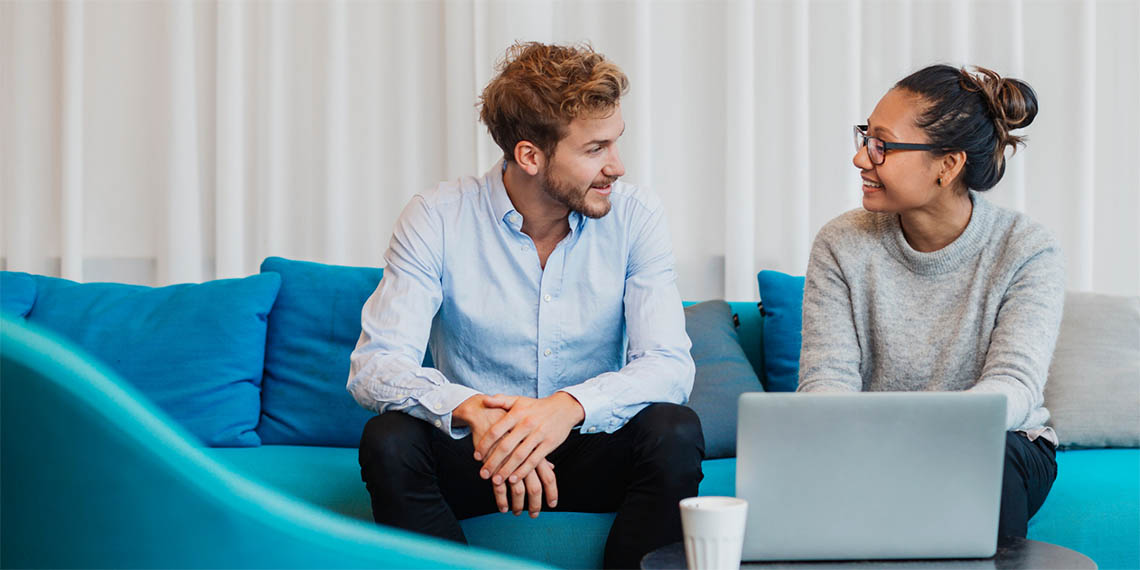 En kvinna och en man samtalar framför en uppslagen bärbar dator