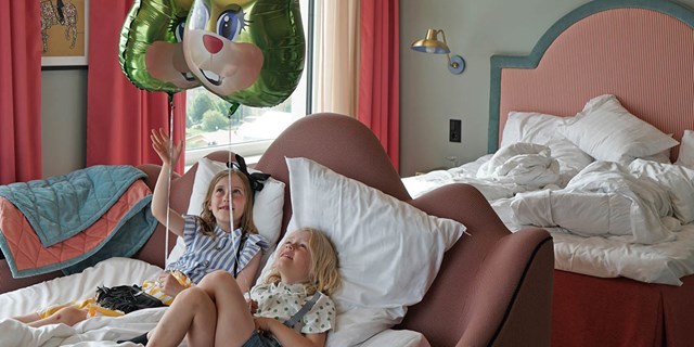 Två barn ligger i en hotellsäng och håller varsin heliumballong i form av den kanin som Liseberg har som sitt signum. .