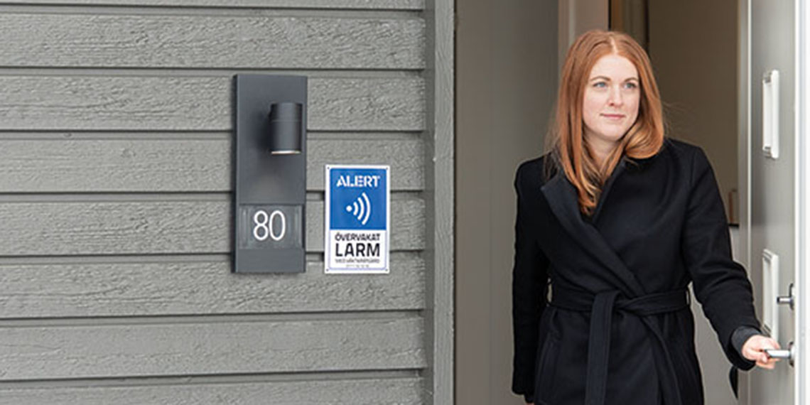 En kvinna öppnar dörren till sitt hus. På husväggen finns en skylt där det står Alert Alarm. 